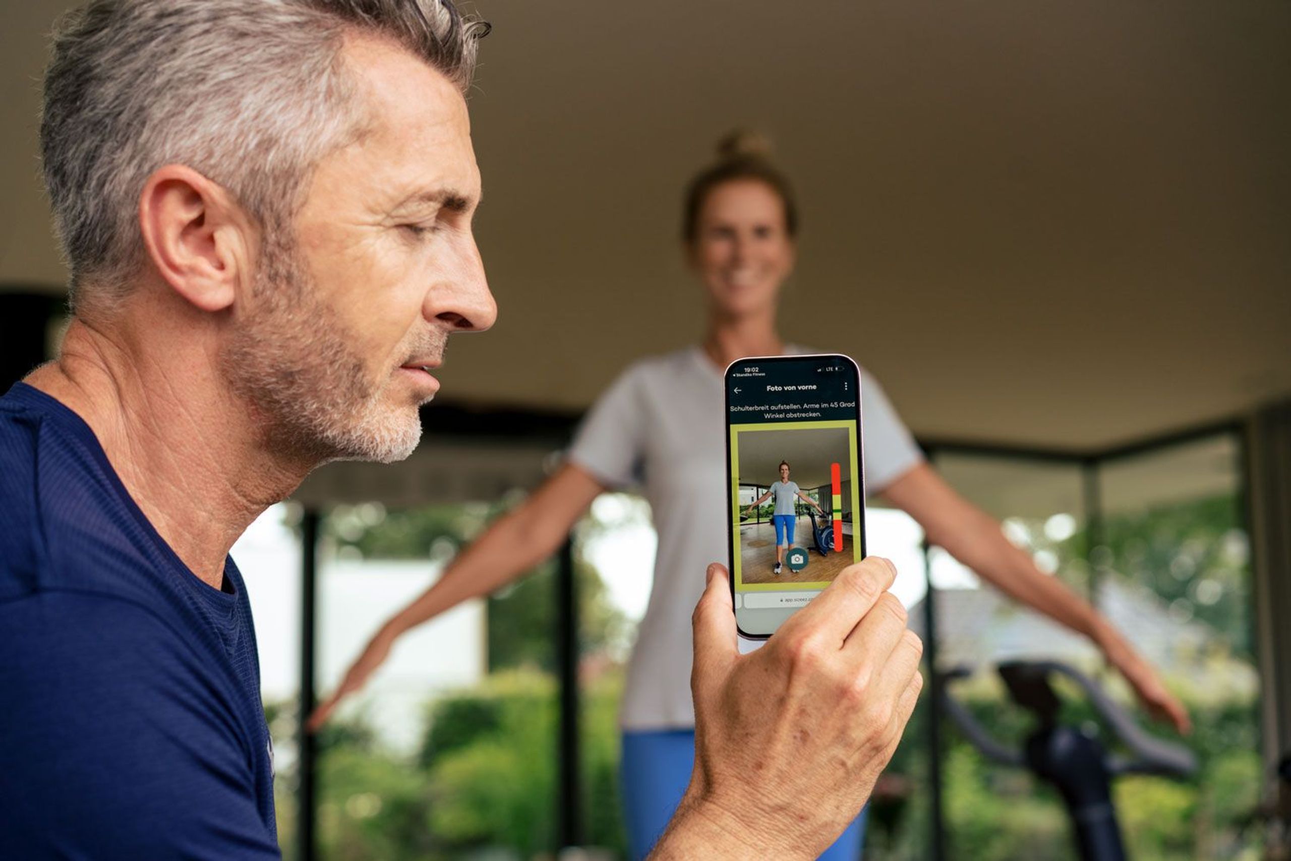 Mann hält Smartphone mit Skandika Fitness App in den Vordergrund, während die App Körpermaße einer Sportlerin ermittelt
