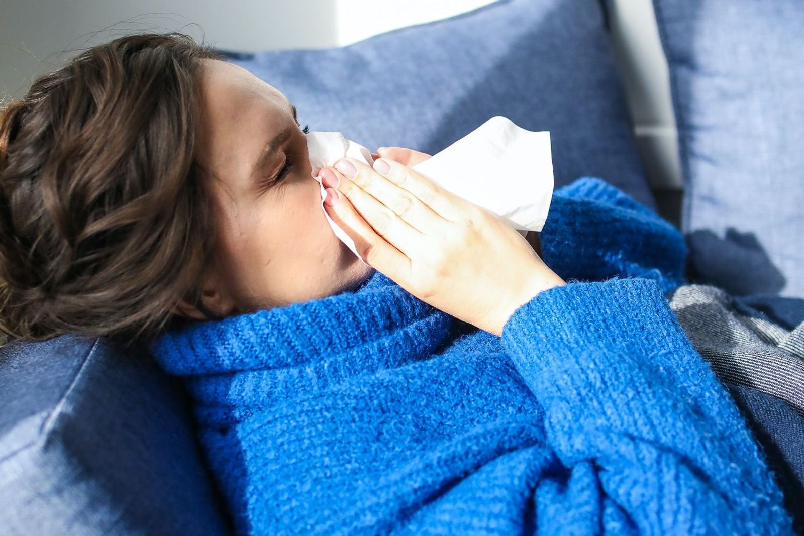 Frau mit blauem Pullover auf der Couch, die sich die Nase putzt