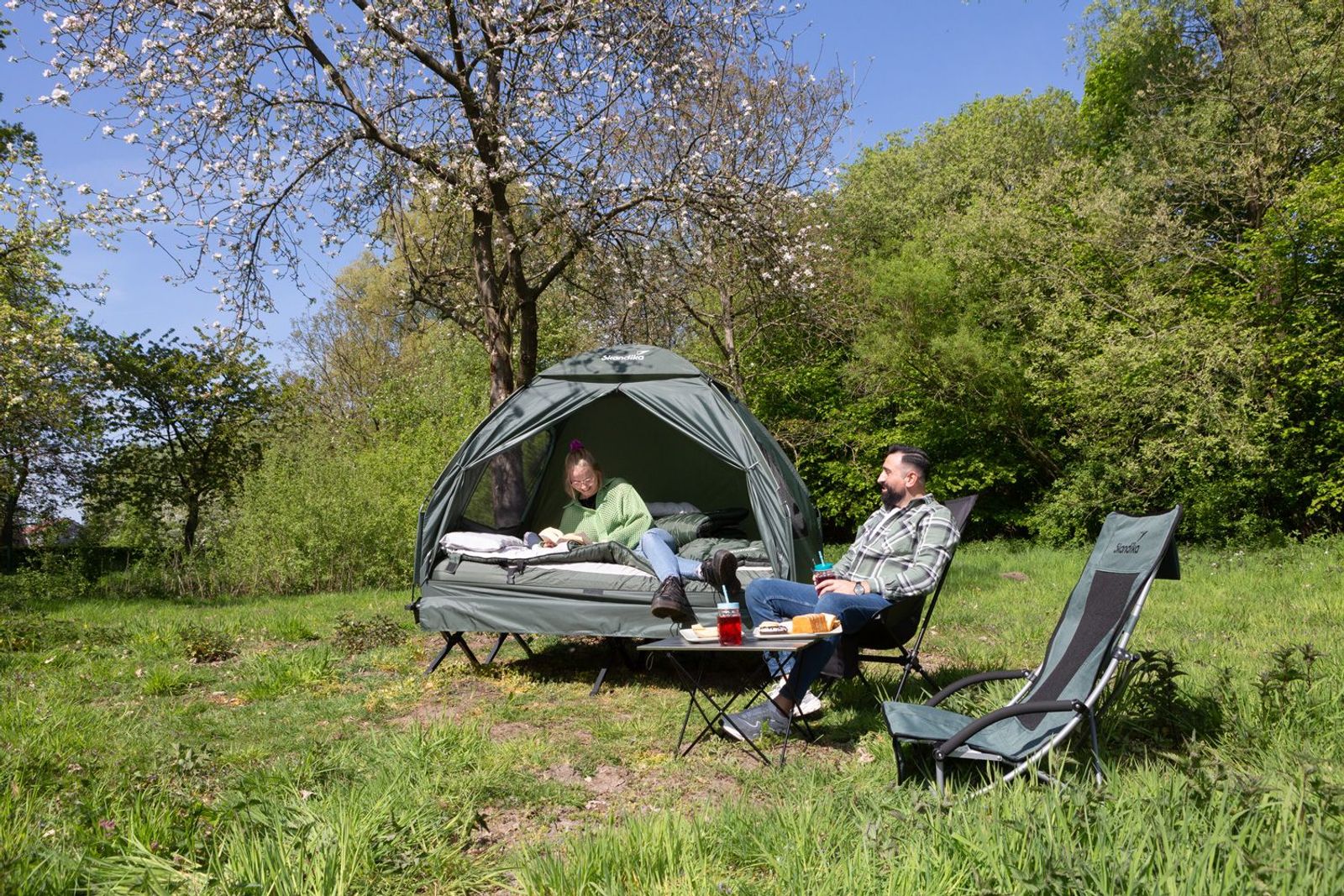 Zeltliege Haug in army green mit zwei Personen, dem Campingtisch Ruka und zwei Campingstühlen in der Natur