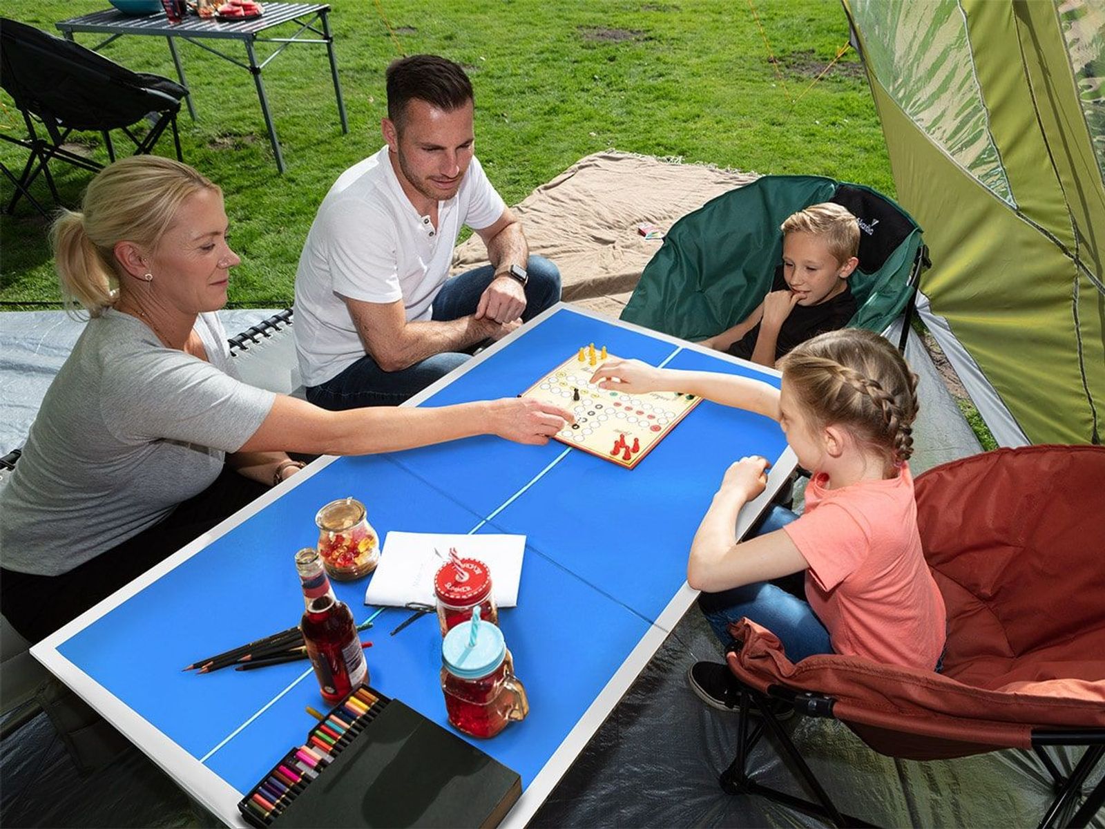 Familie beim Camping mit Multi-Spieletisch, auf dem "Mensch ärger dich nicht" gespielt wird.