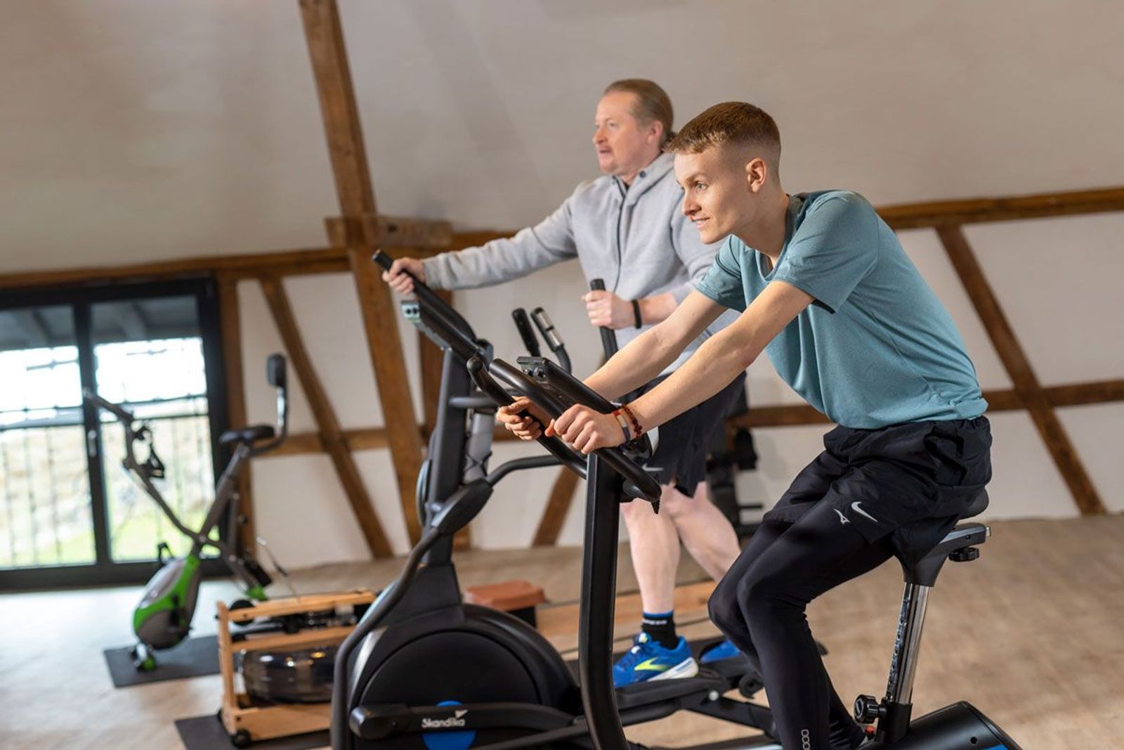 Luke Kelly auf Ergometer mit Joey Kelly im Hintergrund auf einem Ellipsentrainer