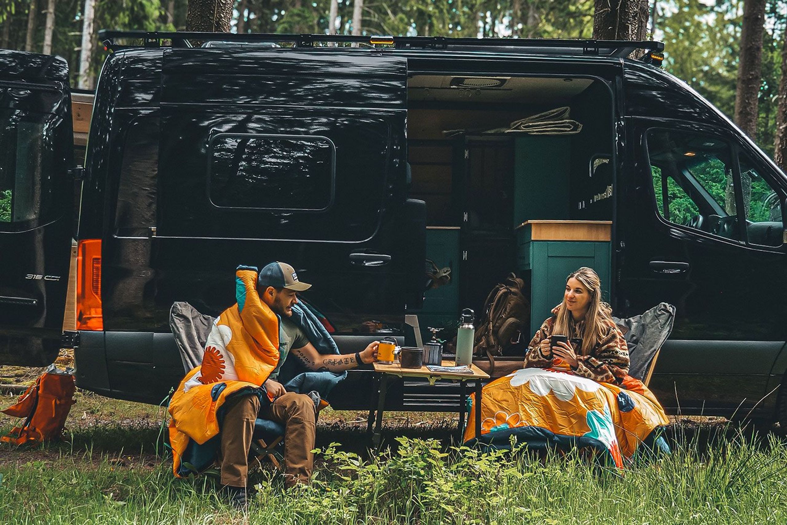 Zwei Camper*innen vor einem ausgebauten Van mit bunten Schlafsäcken auf Stühlen