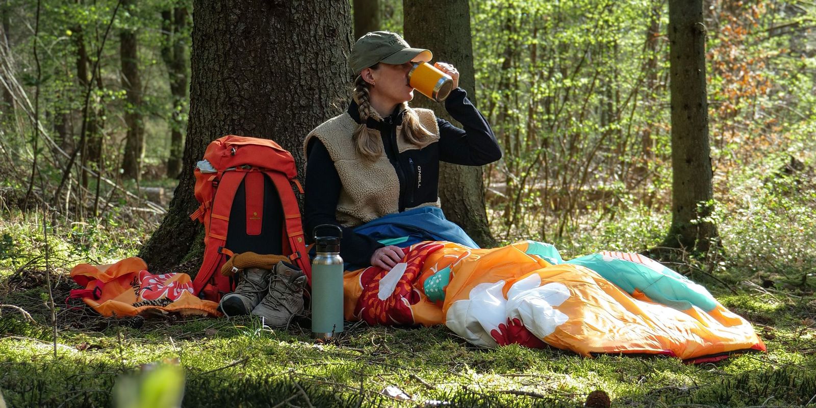 Sovro Schlafsack ausgebreitet auf einem Waldboden mit Frau, die etwas trinkt