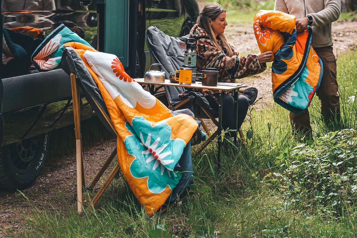 Schlafsack auf einem Stuhl mit zwei Personen im Hintergrund an einem Campervan im Wald
